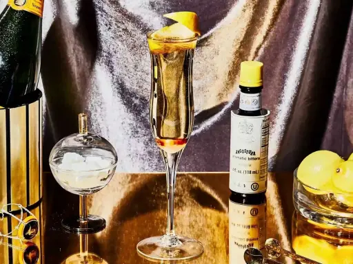 Elegantes Glas mit einem Champagner-Cocktail, garniert mit Zitronenschale. Daneben eine Flasche Angostura, Zuckerwürfel und Zitronen.