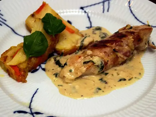 En rullad kyckling insvept i bacon, serverad med en krämig sås till kyckling och gratinerade potatisbitar med gröna blad.