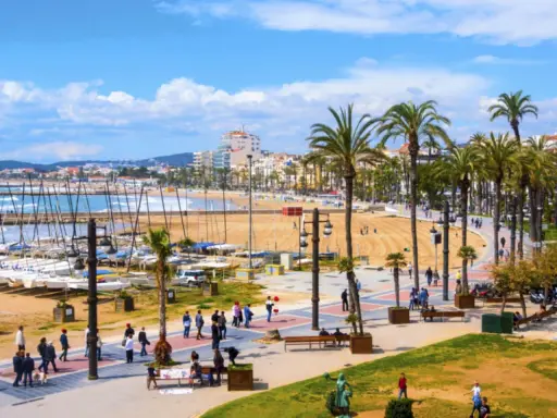 En livlig strandpromenad i Sitges, Spanien, med palmer, en sandstrand, båtar i hamnen och människor som njuter av solen.