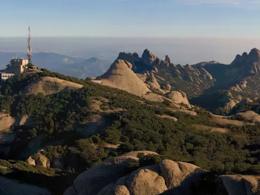 Edificio con antena en la cima de una montaña rocosa, con vistas a un paisaje montañoso y brumoso que se extiende hasta el horizonte, evocando la sensación de estar en planos cerca de Barcelona.