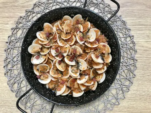 Almejas con jamón en una paellera, con trocitos de jamón y perejil picado, sobre un mantel de madera.