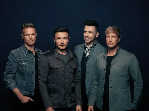 Medlemmar av Westlife poserar för en studiofotografering. De bär moderna jackor och ser direkt in i kameran.