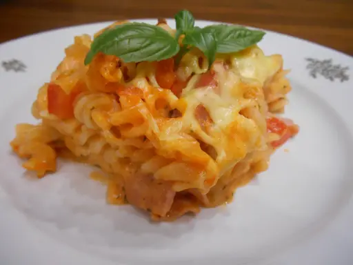 Pyszna zapiekanka z szynką, makaronem fusilli, pomidorami i ciągnącym się serem, ozdobiona listkami bazylii.