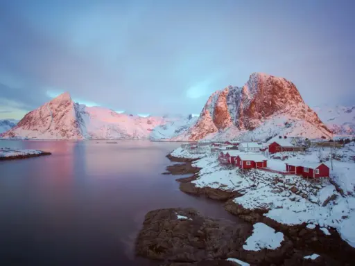 Vackert lofoten vinterlandskap med snöklädda berg, röda hus vid vattnet och en rosa himmel.