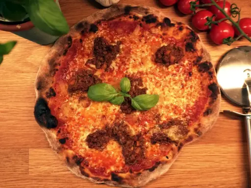 En napolitansk pizza med köttfärs som topping, garnerad med färsk basilika. Bredvid ligger en pizzaskärare och körsbärstomater.