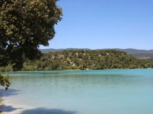 Vue paisible de l'étang de la Bonde, avec ses eaux turquoise bordées de verdure et de collines boisées sous un ciel bleu clair.