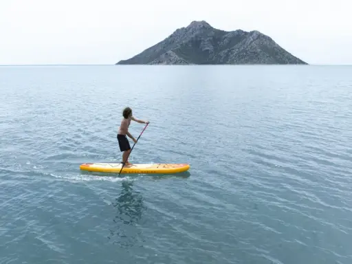 Chłopak na desce sup Aqua Marina przemierza spokojne morze w kierunku skalistej wyspy.