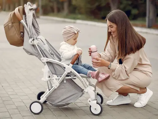 Mama karmi córeczkę z butelki, siedząc obok wózka. Idealne wózki dla dziewczynki na spacer.