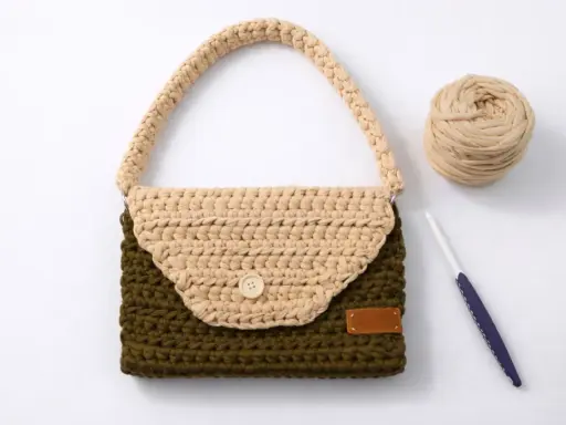 Bolso de trapillo bicolor, verde y beige, con asa y botón de madera. A su lado, ovillo de lana y ganchillo.