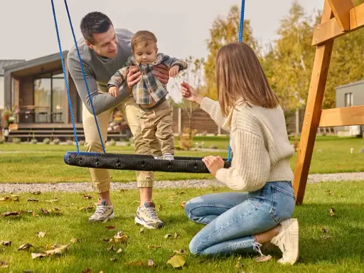 Radosne zabawy w ogrodzie: tata podtrzymuje dziecko na huśtawce, mama zachęca do zabawy.