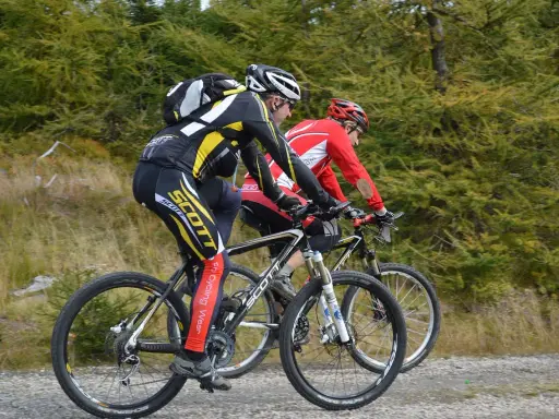 Dwóch kolarzy górskich w kaskach i strojach sportowych jedzie po szutrowej drodze. Jeden z nich, z tyłu, ma na nogawkach napis "Scott". Słychać, jak rower skrzypi podczas pedałowania.