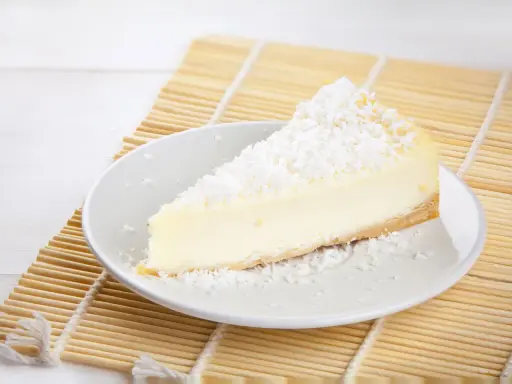Une part de tarte noix de coco, crémeuse et généreusement saupoudrée de noix de coco râpée, repose sur une assiette blanche, prête à être dégustée.