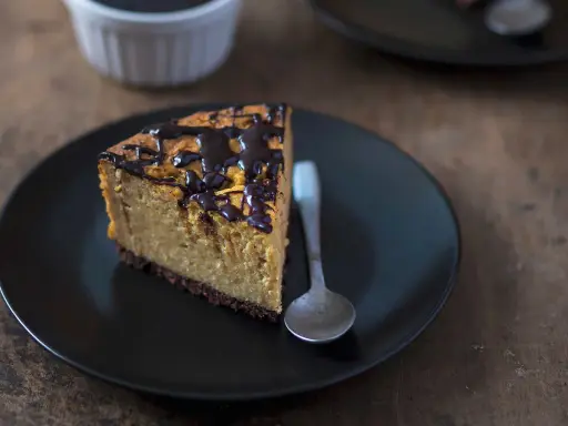 Tranche de cheesecake sans gluten sans lactose, nappée de chocolat noir, sur une assiette noire avec une cuillère.