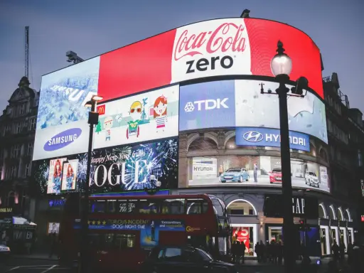 Wielkie ekrany reklamowe na placu, prezentujące różne rodzaje reklamy: Coca-Cola Zero, Samsung, Vogue, Hyundai, TDK. Widać też autobus i ludzi.
