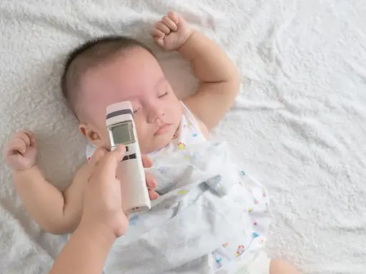 Jak mierzyć temperaturę u niemowlaka termometrem bezdotykowym? Ręka trzyma termometr przy czole śpiącego maluszka.