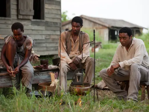 Tre persone sedute all'aperto, in un'ambientazione che ricorda i film biografici sulla schiavitù.