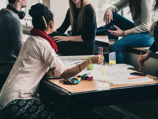 Eine Gruppe junger Leute arbeitet an einem Projekt. Eine Frau mit **Lernschwäche** hält einen Stift und erklärt etwas.