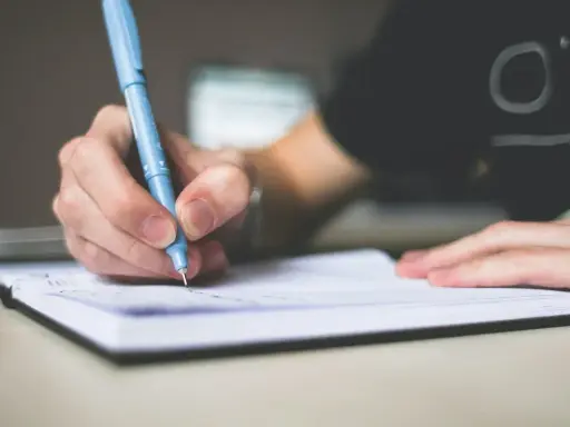 Dłoń pisze niebieskim długopisem w notatniku. To może być początek pisania opinii, esej przykład.