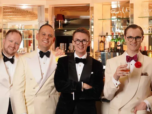 Four men in stylish suits and bow ties, embodying creative black tie, pose at a bar. One holds a cigar, another a drink.