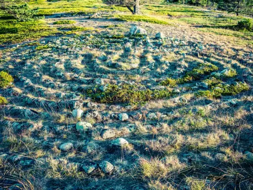 Stenlagd labyrint i Sverige, omgiven av gräs och buskar. Solen kastar långa skuggor över den uråldriga stensättningen.