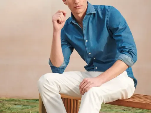 A man poses in a blue denim shirt and crisp white jeans, looking relaxed on a wooden bench.