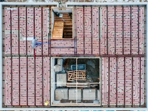 Siatka rur do ogrzewania podłogowego ułożona co 10-15 cm na betonowej płycie fundamentowej. Widoczne schody i materiały budowlane.