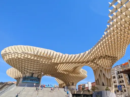 Der Metropol Parasol in Sevilla ragt mit seiner Holzkonstruktion in den blauen Himmel. Menschen steigen die Treppen hinauf.