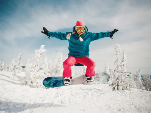Dziewczyna w różowej czapce i goglach skacze na snowboardzie na tle zaśnieżonych drzew. To jest właśnie snowboard!