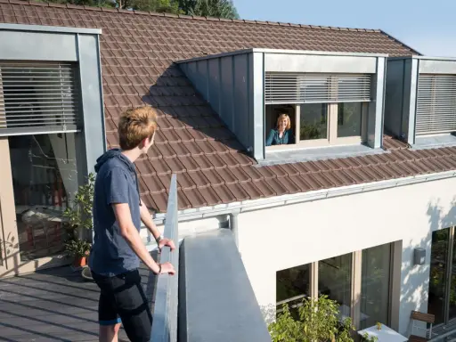 Junge Person auf Balkon blickt zu Frau in Schleppgaube. Verschiedene Dachgauben Arten sind zu sehen.