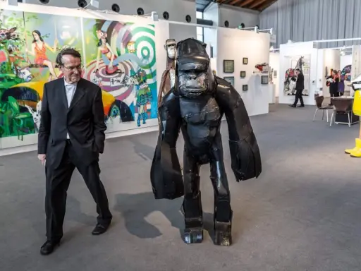Mann in Anzug neben Gorilla-Skulptur auf Kunstmesse. Bunte Gemälde im Hintergrund zeigen aktuelle Kunst.