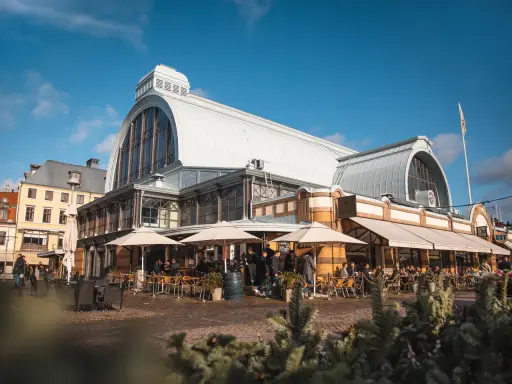 Människor sitter vid bord utomhus framför den imponerande **stora saluhallen** under en klarblå himmel.