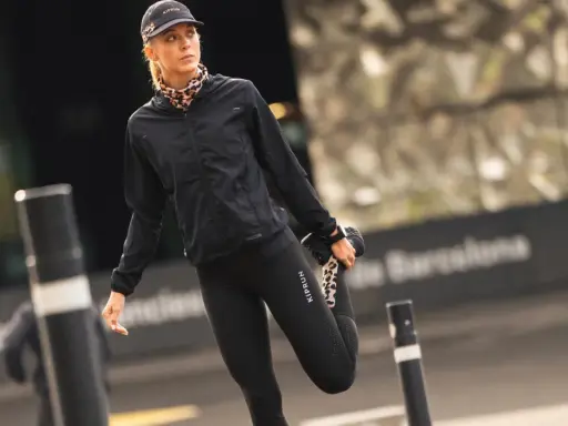 Kobieta w czapce z daszkiem i sportowym stroju rozciąga nogę na tle miejskiego krajobrazu.