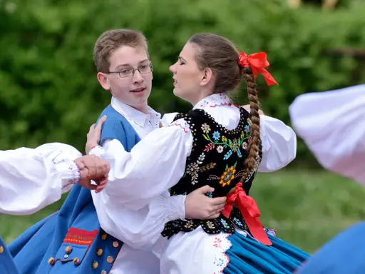 Młoda para w strojach ludowych tańczy mazura. Chłopiec w niebieskiej kamizelce, dziewczyna w haftowanej kamizelce i warkoczu z czerwoną wstążką.