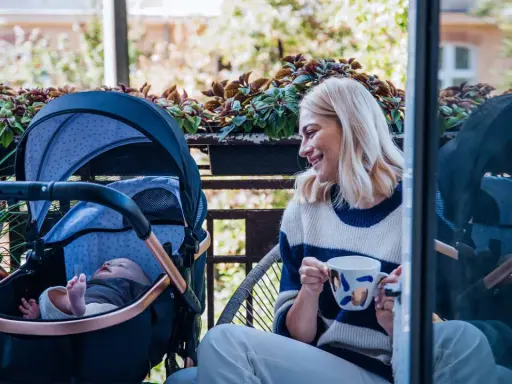 Uśmiechnięta mama z kawą na balkonie, obok wózek z dobrą amortyzacją i śpiące dziecko.