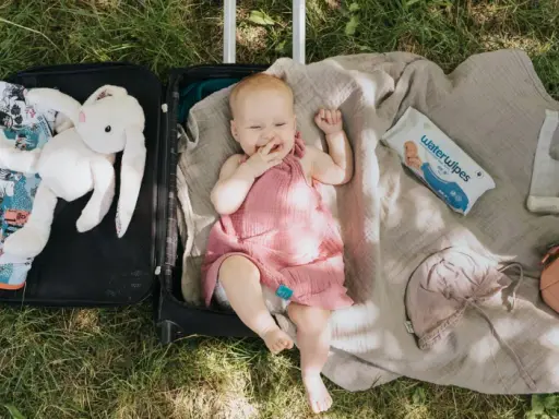 Uśmiechnięte niemowlę w walizce, gotowe na podróże z niemowlakiem. Obok pluszowy króliczek, kocyk, kosmetyczka i chusteczki.