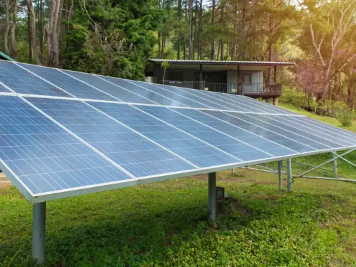 Grandes panneaux solaires au sol, installés dans un jardin verdoyant, captent l'énergie du soleil pour alimenter une maisonnette en bois.