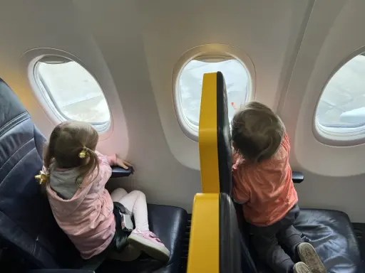 Dzieci patrzą przez okno samolotu Wizz Air. Dziewczynka w różowej bluzie i chłopiec w pomarańczowej koszulce, siedzą obok siebie.