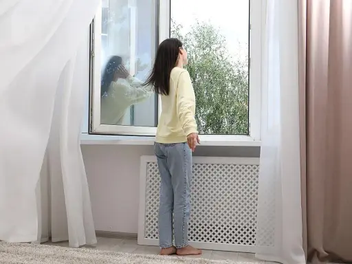 Mujer abre ventana para ventilar la casa en invierno. Reflejo en el cristal muestra a otra persona.