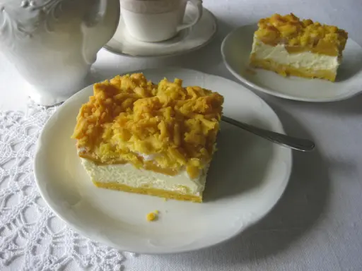 Sernik z brzoskwiniami na białym talerzyku, z kruszonką na wierzchu. Obok filiżanka i dzbanek.