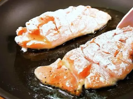 Due filetti di petto di pollo infarinati cuociono in padella, sfrigolando nell'olio caldo.