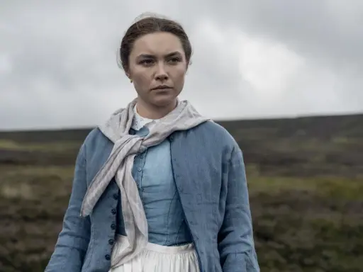 Florence Pugh i en blå klänning och jacka, med en scarf runt halsen, står i ett öppet landskap. En scen från filmer med Florence Pugh.