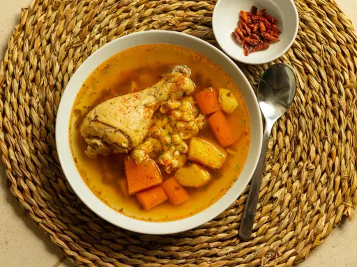 Un confortante brodo di pollo con pezzi di pollo, carote e patate, servito su una tovaglietta di paglia.