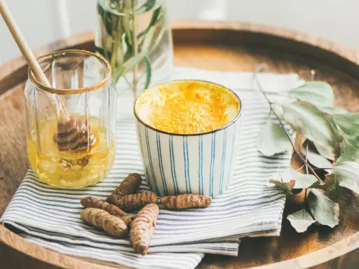 Aromaticzne złote mleko w kubeczku, obok słoiczek z miodem i korzenie kurkumy. Idealne na chłodne wieczory.
