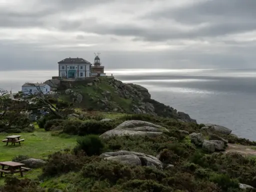 Leuchtturm und Gebäude auf felsiger Küste bei Finisterre, Spanien. Grauer Himmel über dem Meer.