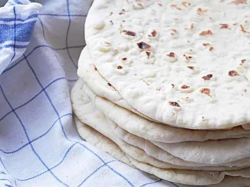 En hög med nybakade tortillabröd, perfekta för ditt nästa tortilla recept. Bredvid ligger en blåvitrutig kökshandduk.