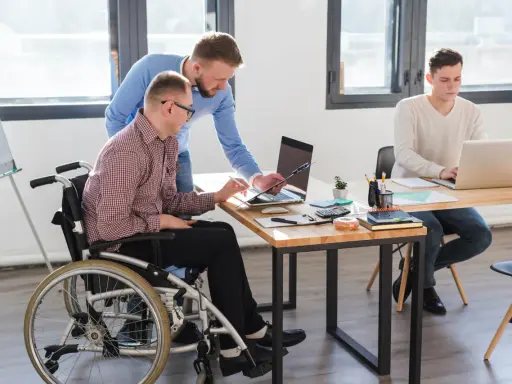 Współpraca w biurze: mężczyzna na wózku inwalidzkim, z umiarkowanym stopniem niepełnosprawności, pracuje z kolegą przy laptopie.