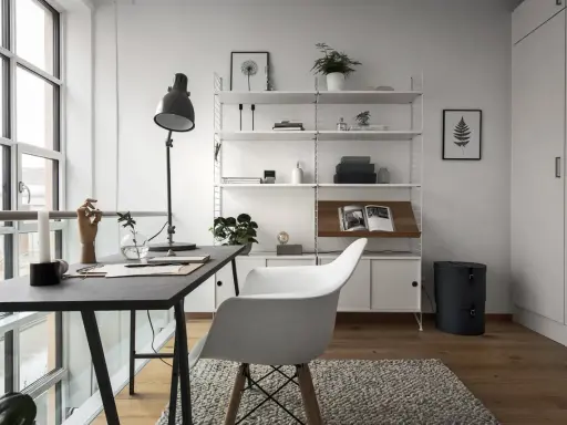 Ett modernt hemmakontor med ett mörkt skrivbord, en vit stol och en stilren, inredd bokhylla. Ljuset flödar in från stora fönster.