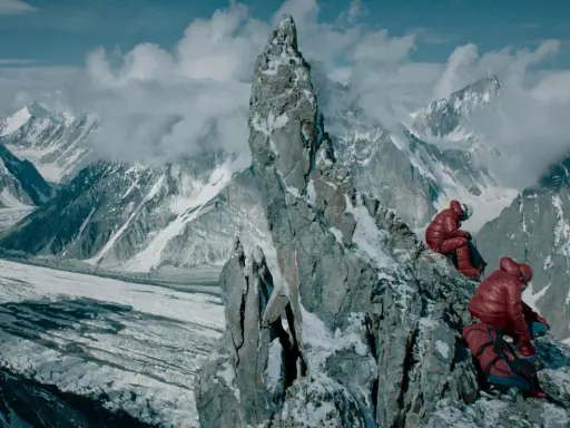 Dwóch alpinistów w czerwonych kurtkach odpoczywa na skalistej grani, podziwiając rozległy widok na lodowiec i ośnieżone szczyty. Broad peak góra w oddali.