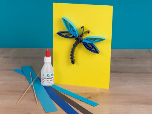 Eine Libelle aus Papierstreifen, erstellt mit einer Quilling-Anleitung. Kleber und bunte Streifen liegen bereit.