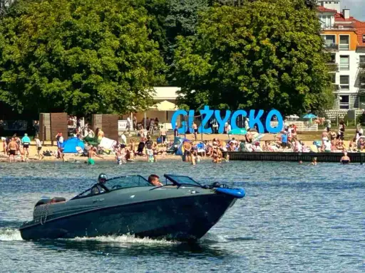 Motorówka sunie po wodzie, w tle plaża pełna ludzi i wielki napis "GIŻYCKO". Idealny dzień na Mazurach.